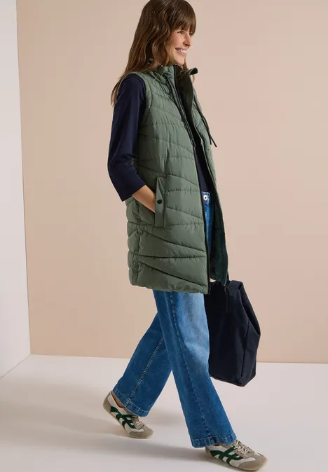 Lange Steppweste mit Stehkragen shadow khaki Frau mit langer, gesteppter Kapuzen-Weste, dunklem Oberteil, weitem Jeans und Sneakers, trägt Shopper-Tasche.