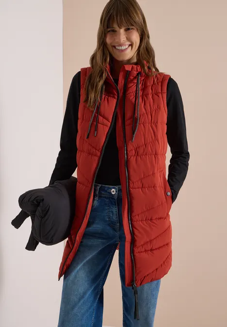 Lange Steppweste mit Stehkragen light russet red Frau mit roter Steppweste, schwarzem Top und Jeans, hält eine schwarze Jacke und lächelt in die Kamera.