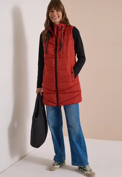 Lange Steppweste mit Stehkragen light russet red Frau lächelt, trägt rote Steppweste mit Kapuze, schwarzen Pullover, blaue Jeans und schwarze Tasche.