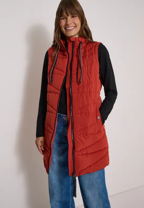 Lange Steppweste mit Stehkragen light russet red Frau in roter, langer Steppweste mit Kapuze, schwarzem Shirt und weiten Jeans, lächelnd vor heller Wand.