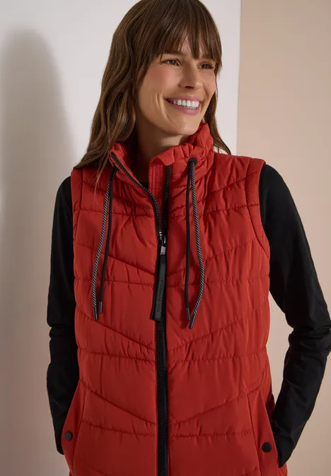 Lange Steppweste mit Stehkragen light russet red Frau lächelt und trägt rote, gesteppte Weste mit Kapuze und schwarzen Langarmshirt.