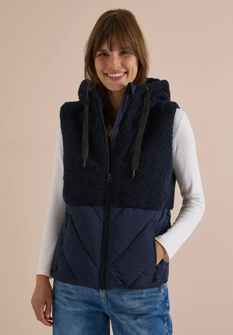 Teddy-Weste mit Steppteil und Zipper dark navy blue Frau trägt eine dunkelblaue, ärmellose Weste mit Fleece-Kapuze, weißes Langarmshirt und Jeans, lächelt entspannt.