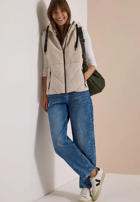 Eine junge Frau lehnt lächelnd an einer Wand, trägt Jeans, Weste, Sneakers und Rucksack.