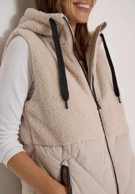 Eine Frau trägt eine beige Teddy-Fleece-Weste mit Kapuze und Reißverschlusstaschen.