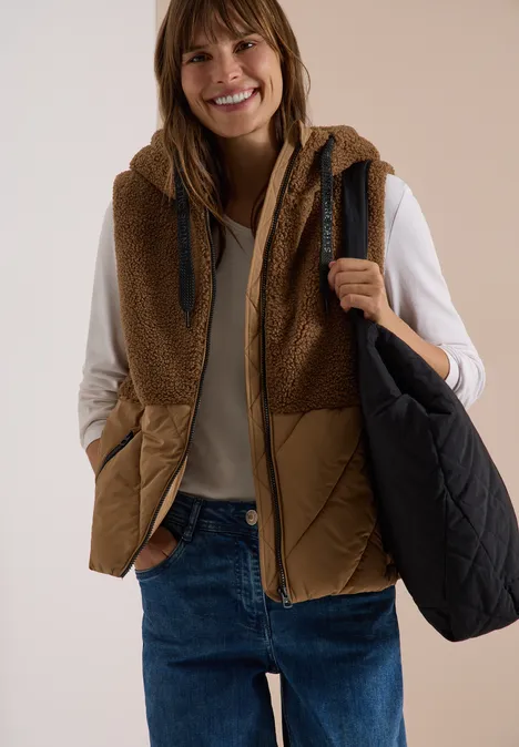 Teddy-Weste mit Steppteil und Zipper dark toffee brown Lächelnde Frau in brauner Fleece-Steppweste, beigem Pullover, Jeans und schwarzer Tasche vor hellem Hintergrund.