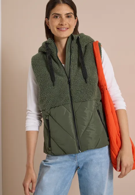 Teddy-Weste mit Steppteil und Zipper shadow khaki Frau trägt olivgrüne, ärmellose Kapuzenweste mit Stepp-Details, weißes Langarmshirt und orangene Einkaufstasche.