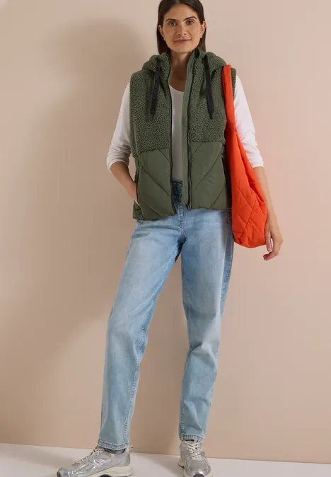 Teddy-Weste mit Steppteil und Zipper shadow khaki Frau trägt khakigrüne Steppweste über weißem Shirt, Jeans und Sneakern, hält orange Tasche vor beiger Wand.