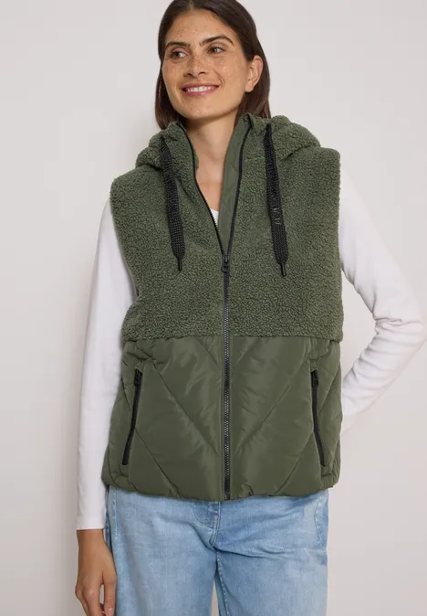 Teddy-Weste mit Steppteil und Zipper shadow khaki Frau trägt weißes Langarmshirt, Jeans und eine grüne, ärmellose Fleeceweste mit Kapuze und Reißverschluss.
