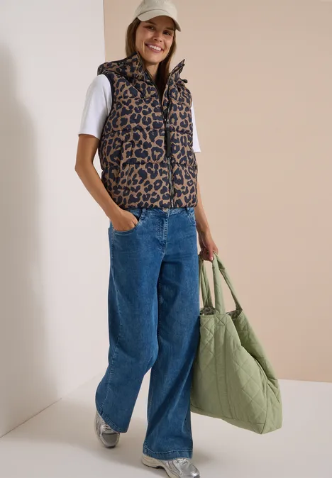 Steppweste mit Kapuze und Taschen creamy mocha Frau in leo-gemusterter Weste, weißem Shirt, weitem Jeans, Sneakern und großer grüner Tasche.