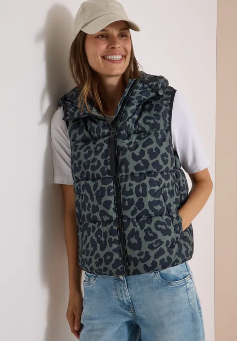 Steppweste mit Kapuze und Taschen dried olive green Junge Frau trägt Jeans, weißes T-Shirt, Leopard-Weste und Baseballcap, lächelt.