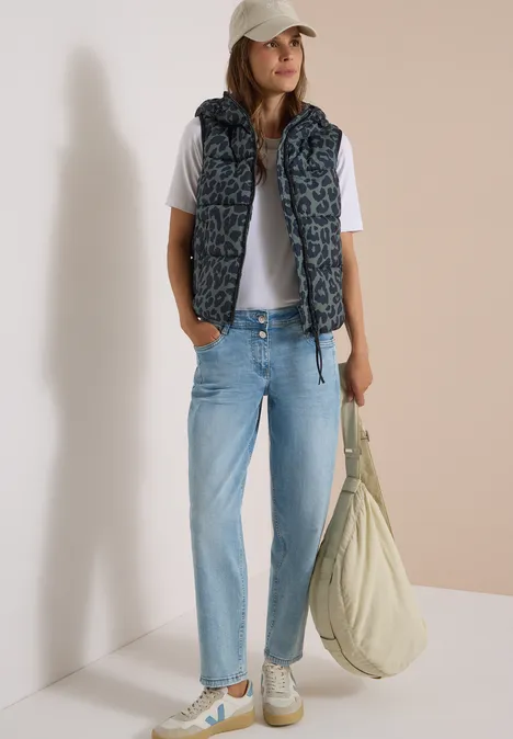Steppweste mit Kapuze und Taschen dried olive green Frau in Jeans, weißem T-Shirt, gemusterter Daunenweste und Cap, mit großer Tasche vor neutralem Hintergrund