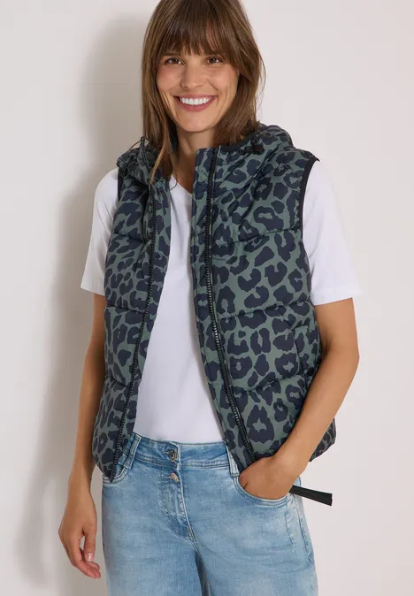 Steppweste mit Kapuze und Taschen dried olive green Frau mit gemusterter Kapuzenweste über weißem T-Shirt und Jeans, lächelnd.