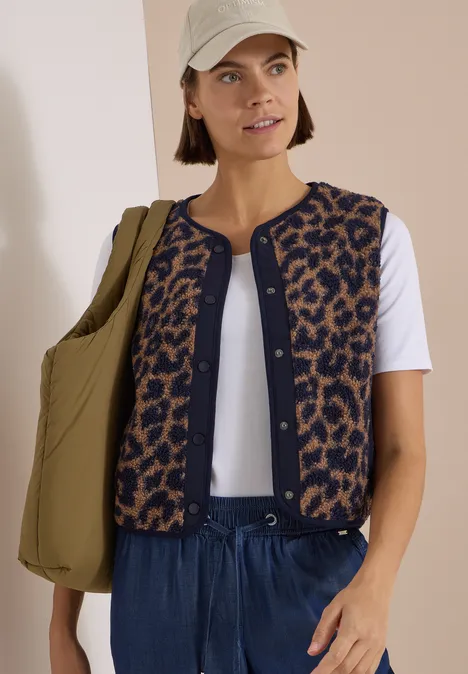 Teddy-Weste mit Softshelldetails und Leo-Muster creamy mocha Frau mit beiger Cap, weißem Shirt, kurzer Leo-Weste, blauen Hosen und olivgrüner Tasche.