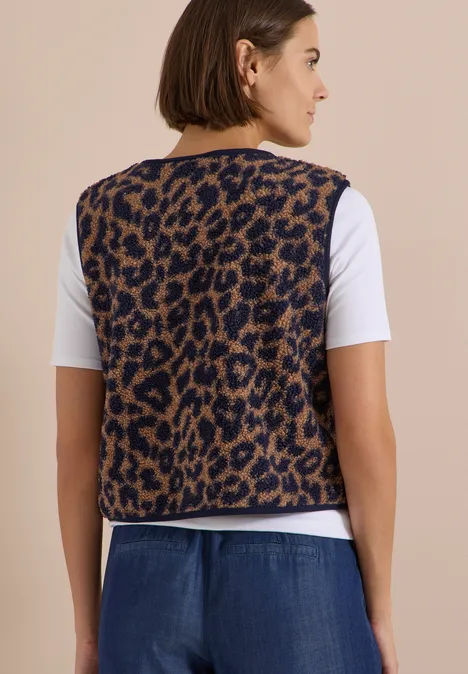 Teddy-Weste mit Softshelldetails und Leo-Muster creamy mocha Frau seitlich in weißem T-Shirt, braun-gemusterter Leoparden-Weste und Jeanshose vor beigem Hintergrund.