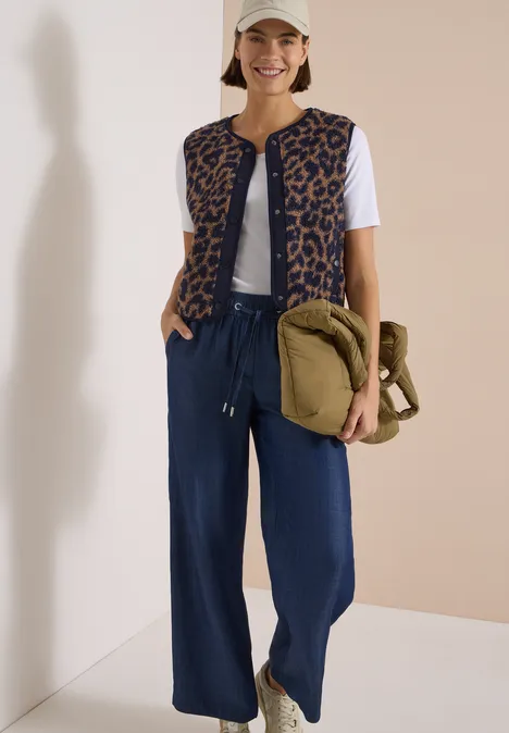 Teddy-Weste mit Softshelldetails und Leo-Muster creamy mocha Frau im Freizeitlook mit Cap, weißem T-Shirt, Leo-Weste, weiten Jeanshosen und grüner Umhängetasche.