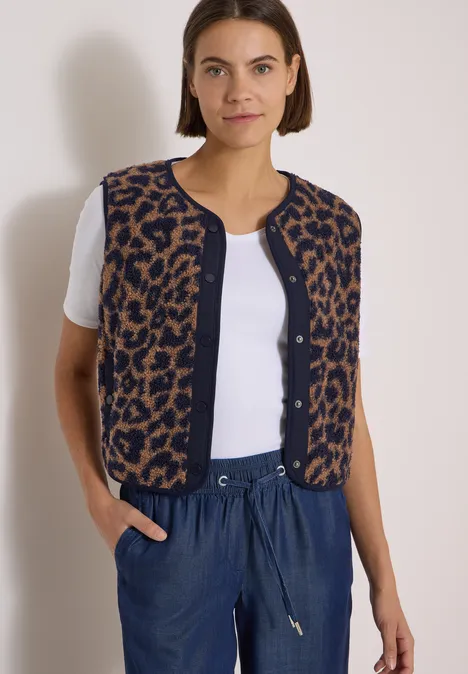 Teddy-Weste mit Softshelldetails und Leo-Muster creamy mocha Frau trägt weißes Shirt, leo-Strickweste ohne Ärmel und blaue Hose vor hellgrauem Hintergrund.