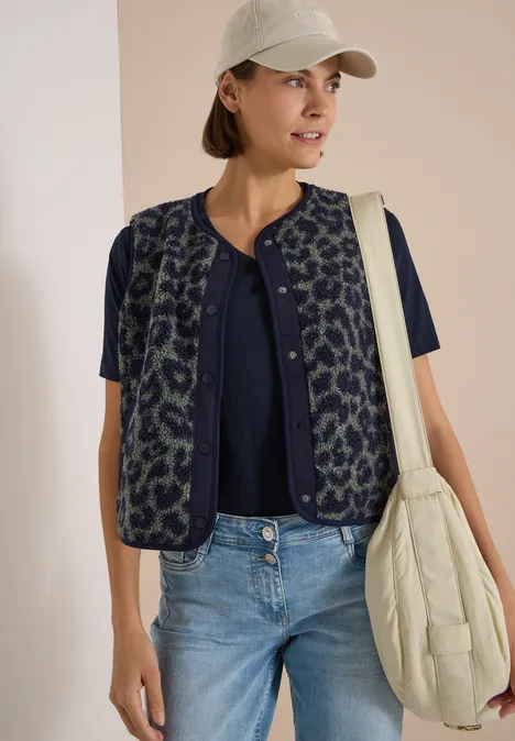 Teddy-Weste mit Softshelldetails und Leo-Muster dried olive green Frau mit weißem Cap, dunkelblauer Bluse, gemusterter Weste, Jeans und cremefarbener Stepp-Tasche.