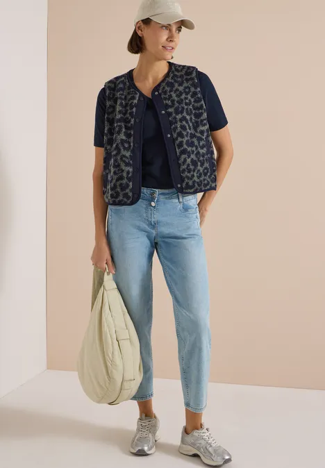 Teddy-Weste mit Softshelldetails und Leo-Muster dried olive green Frau in weißer Kappe trägt gestepste Weste über dunkelblauem Shirt, hellblaue Jeans, graue Sneaker und große Tasche.