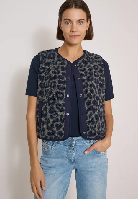Teddy-Weste mit Softshelldetails und Leo-Muster dried olive green Frau in blauem T-Shirt mit grüner, leo-gemusterter Weste und hellen Jeans vor weißem Hintergrund.