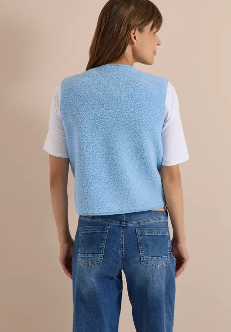 Kobieta widziana od tyłu w jasnoniebieskiej polarowej kamizelce, białym T-shircie i niebieskich dżinsach na beżowym tle.