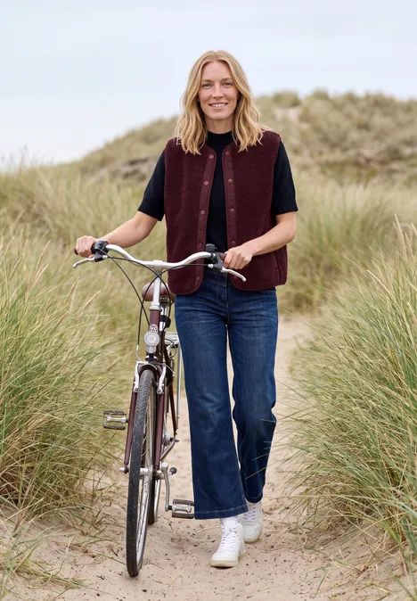 Teddy-Weste mit Softshelldetails red wine Frau schiebt Fahrrad auf Dünenpfad zwischen hohem Gras am Strand.