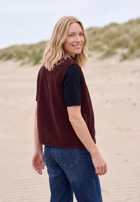 Teddy-Weste mit Softshelldetails red wine Eine lächelnde Frau in einer bordeauxfarbenen Strickweste und Jeans steht am Strand und blickt über die Schulter.