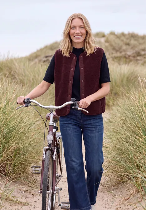 Teddy-Weste mit Softshelldetails red wine Eine lächelnde Frau geht mit einem Fahrrad durch grasbewachsene Dünen am Strand.