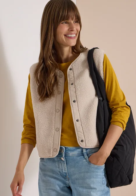 Femme souriante en pull jaune, gilet en sherpa beige, jean et sac noir.