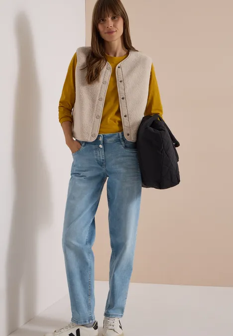 Femme en pull moutarde, gilet beige et jean bleu clair, portant un sac foncé devant un mur neutre.