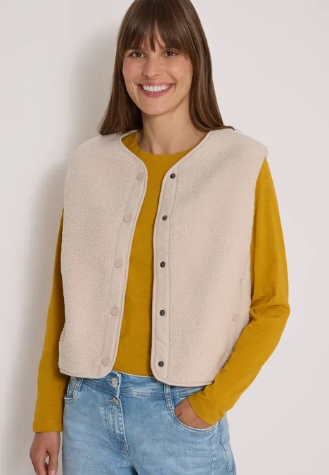 Femme souriante en t-shirt jaune et gilet beige, jean décontracté, mains dans les poches.