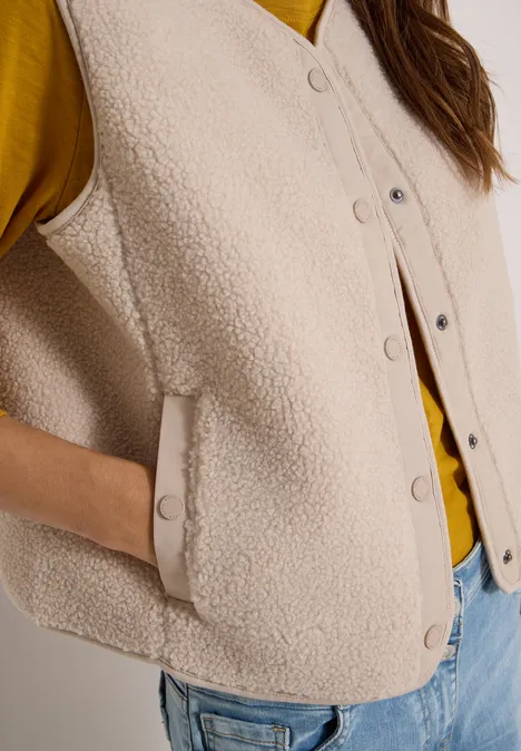 Gros plan d'un gilet en sherpa beige à boutons pression et poche, porté sur un pull jaune.