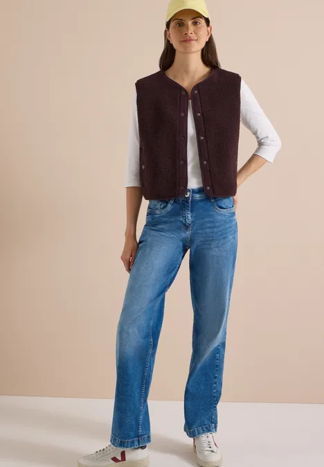 Kobieta w żółtej czapce, brązowej kamizelce, białej koszulce 3/4, niebieskich dżinsach i białych trampkach na neutralnym tle.