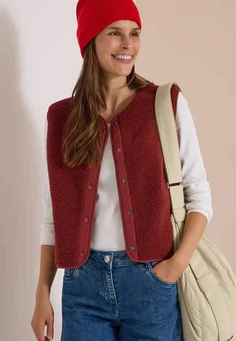 Eine junge Frau trägt rote Weste und Mütze, ein weißes Shirt, Jeans und eine Cremetasche, sie lächelt.