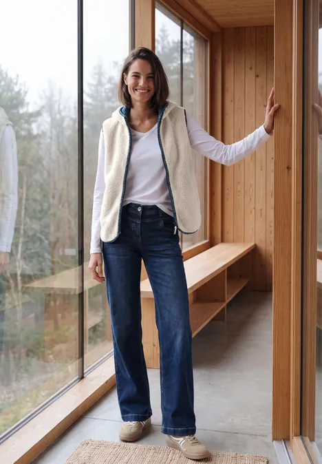 Teddy-Weste mit Kapuze und Zipper teddy vanilla white Eine Frau in weißem Top, Fleeceweste, weiten Jeans und Sneakern steht lächelnd an einem großen Fenster in einem Holzhaus.