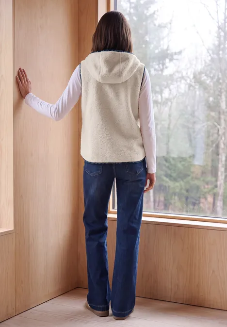 Teddy-Weste mit Kapuze und Zipper teddy vanilla white Person in Fleece-Weste und Jeans steht in einem Holzraum vor grossem Fenster und blickt in den Wald.