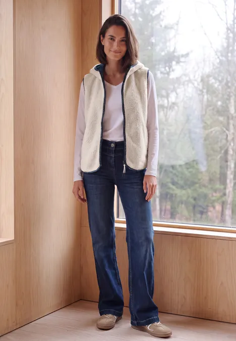 Teddy-Weste mit Kapuze und Zipper teddy vanilla white Eine Frau steht lässig in heller Fleecejacke und Jeans vor einem großen Fenster mit Blick auf den Wald.