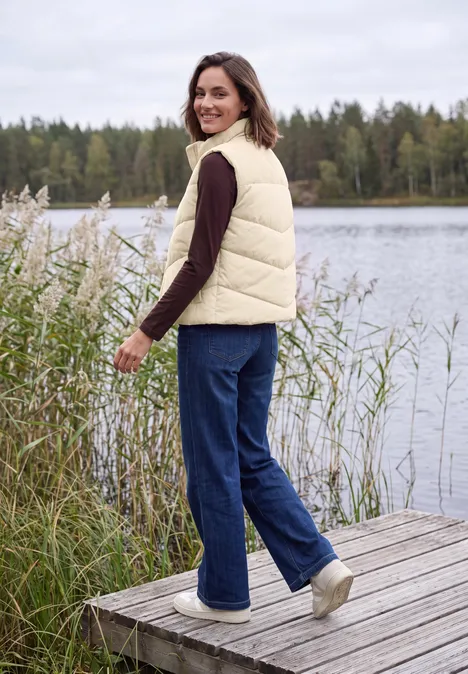 Cord-Weste mit Steppmuster und Zipper pearl beige Frau im cremefarbenen Steppweste und Jeans steht auf einem Holzsteg am See, schaut lächelnd zurück.