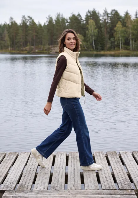 Cord-Weste mit Steppmuster und Zipper pearl beige Frau in Jeans und Weste geht lächelnd über einen Holzsteg am ruhigen See mit Wald im Hintergrund.