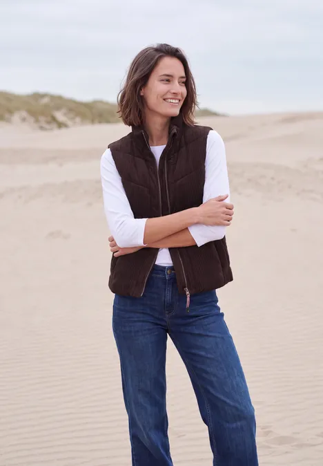 Corduroyvest met doorgestikt patroon en ritssluiting berry red Een vrouw lacht op het strand met een bruin gilet, wit shirt en blauwe jeans voor zandduinen.
