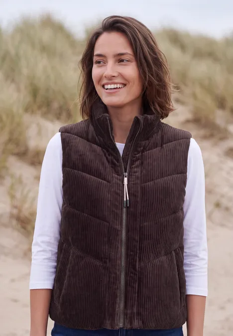 Corduroyvest met doorgestikt patroon en ritssluiting berry red Een vrouw glimlacht bij zandduinen aan zee, ze draagt een bruine corduroy bodywarmer en een wit shirt met lange mouwen.