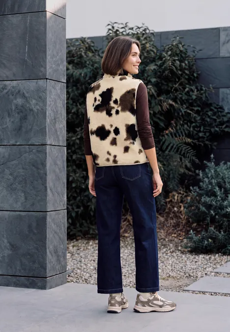 Gilet en peluche à col rond et imprimé pearl beige Frau steht vor grauer Wand, trägt schwarze Hose und Fellweste mit schwarz-weißen Flecken.