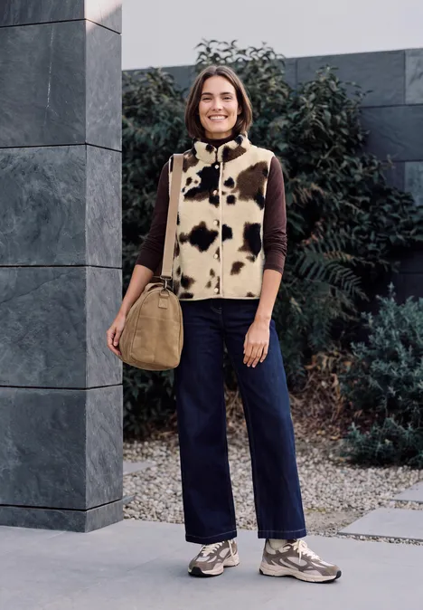 Gilet en peluche à col rond et imprimé pearl beige Frau steht vor modernem Gebäude mit Kuhflecken-Weste, braunem Pullover, blauen Hosen, Sneakers und Tasche.