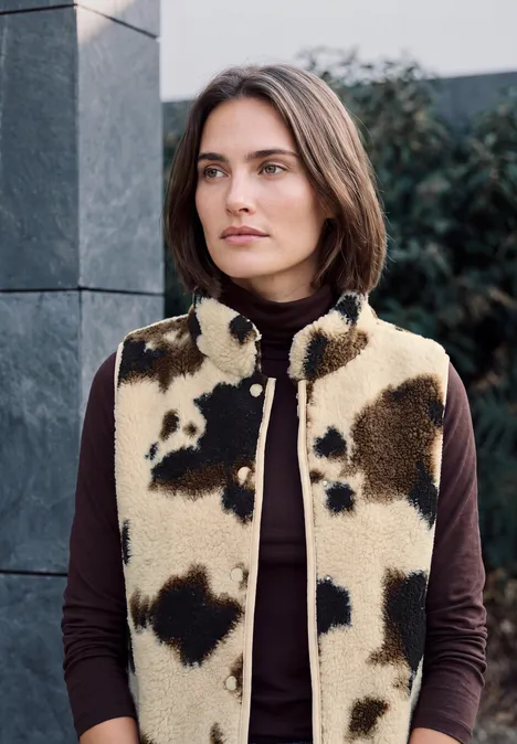 Gilet en peluche à col rond et imprimé pearl beige Frau mit kurzem Bob trägt beige-braune Kuhfell-Weste über dunklem Rollkragenpullover vor moderner Fassade.
