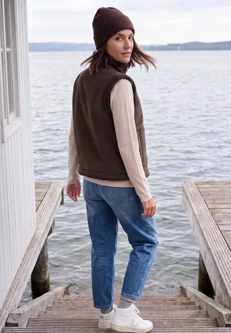 Frau mit brauner Mütze und Weste, Jeans und Sneakern auf einem Bootssteg am Wasser, blickt zurück.