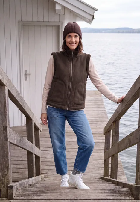 Eine Frau in einer Weste, Jeans und Mütze steht lächelnd auf einem Steg am See.