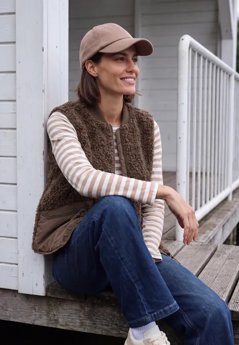 Een glimlachende vrouw zit op een veranda, draagt een bruine fleecebodywarmer, gestreept shirt, jeans en pet.