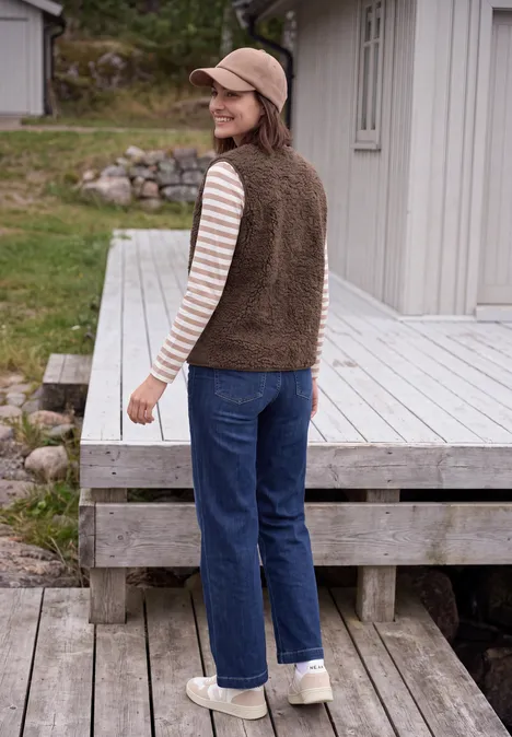 Een lachende vrouw in spijkerbroek, gestreept shirt en fleecevest staat op een houten terras.