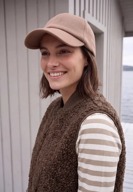 Lachende vrouw met pet, bruine bodywarmer en gestreept shirt voor een houten huis aan het water.