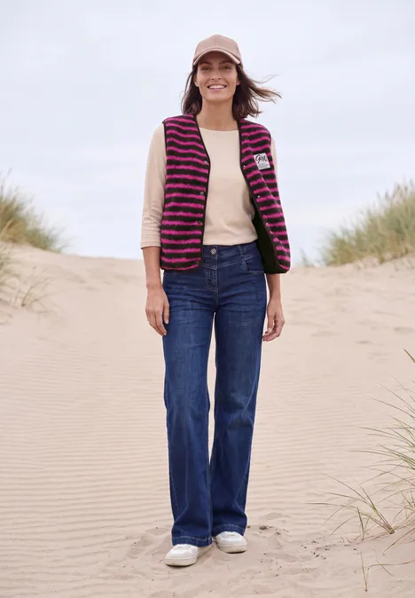 Eine junge Frau steht lächelnd barfuß im Sand, trägt Jeans, beiges Shirt, gestreifte Weste und Cap.