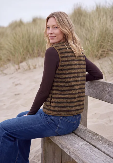 Eine Frau mit gestreiftem Strickweste sitzt auf einer Holzbank am Strand und lächelt.